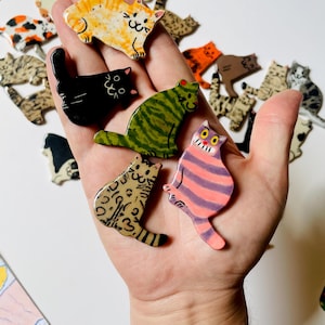 May include: A collection of small, flat, cat-shaped ceramic pins featuring various colors and patterns, including orange tabby, solid black, green striped, leopard print, and a pink and purple striped cat with a wide, toothy grin.