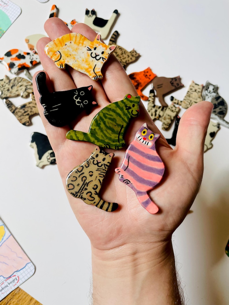 May include: A collection of small, flat, cat-shaped ceramic pins featuring various colors and patterns, including orange tabby, solid black, green striped, leopard print, and a pink and purple striped cat with a wide, toothy grin.