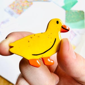 May include: A yellow duck-shaped pin with orange feet and beak, and a black curved line for a smile. The pin has small brown speckles and is held in a person's hand.