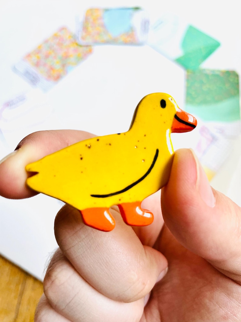 May include: A yellow duck-shaped pin with orange feet and beak, and a black curved line for a smile. The pin has small brown speckles and is held in a person's hand.