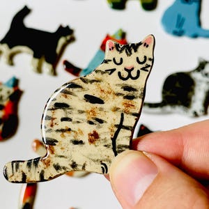 May include: A small, flat, ceramic cat-shaped pin with a cream-colored base, featuring hand-painted black and brown tabby stripes, a pink nose, and closed eyes. The surface has a glossy finish, and other similar cat pins are visible in the background.