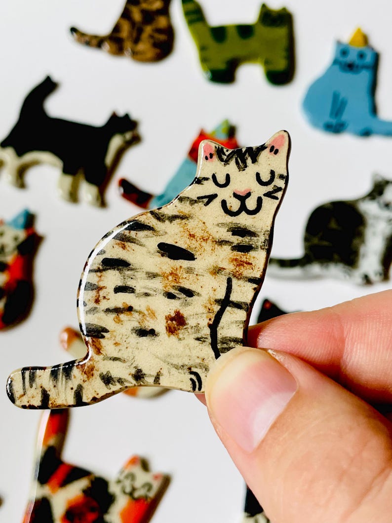 May include: A small, flat, ceramic cat-shaped pin with a cream-colored base, featuring hand-painted black and brown tabby stripes, a pink nose, and closed eyes. The surface has a glossy finish, and other similar cat pins are visible in the background.