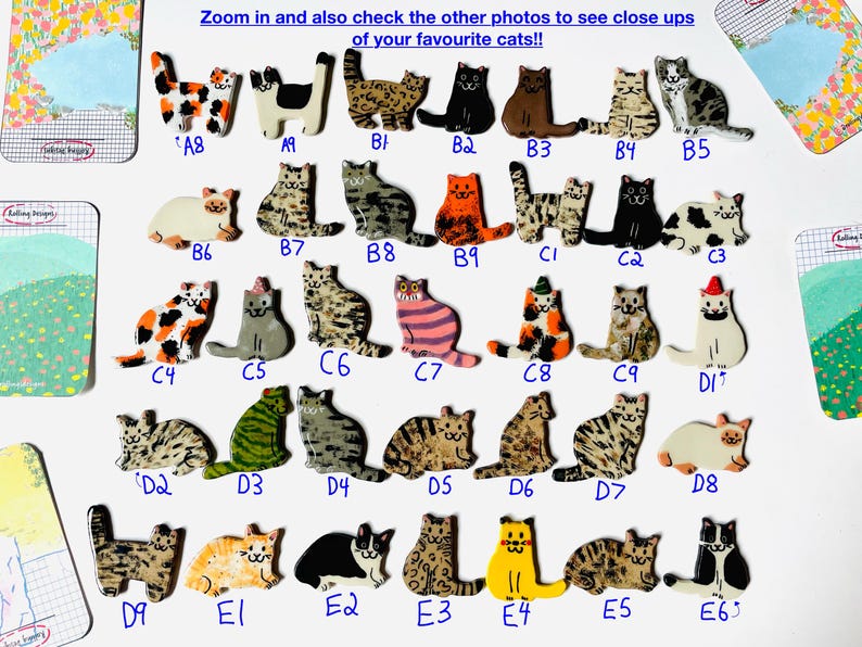 May include: A collection of small, flat, cat-shaped ceramic pins or ornaments, each labeled with a unique alphanumeric code. The cats feature various colors, patterns, and markings, including solid black, orange, tabby, calico, and spotted designs.