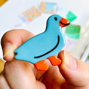 May include: A blue bird-shaped decorative item with orange feet and beak. The bird has a black curved line on its side and a black dot for an eye. The item is held in a person's hand.
