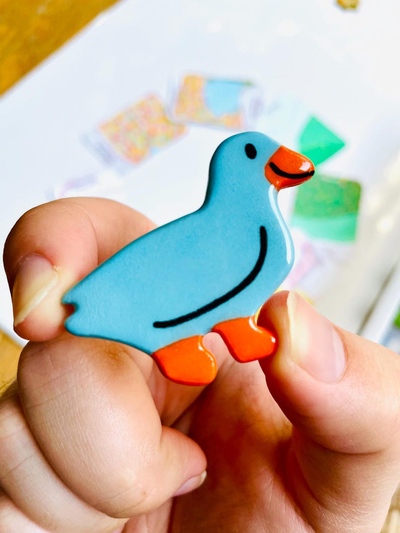 May include: A blue bird-shaped decorative item with orange feet and beak. The bird has a black curved line on its side and a black dot for an eye. The item is held in a person's hand.