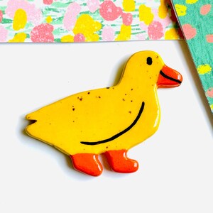 May include: A yellow ceramic duck figurine with orange feet and beak. The duck has a black smiling line and small black eyes. The background includes colorful patterned paper.