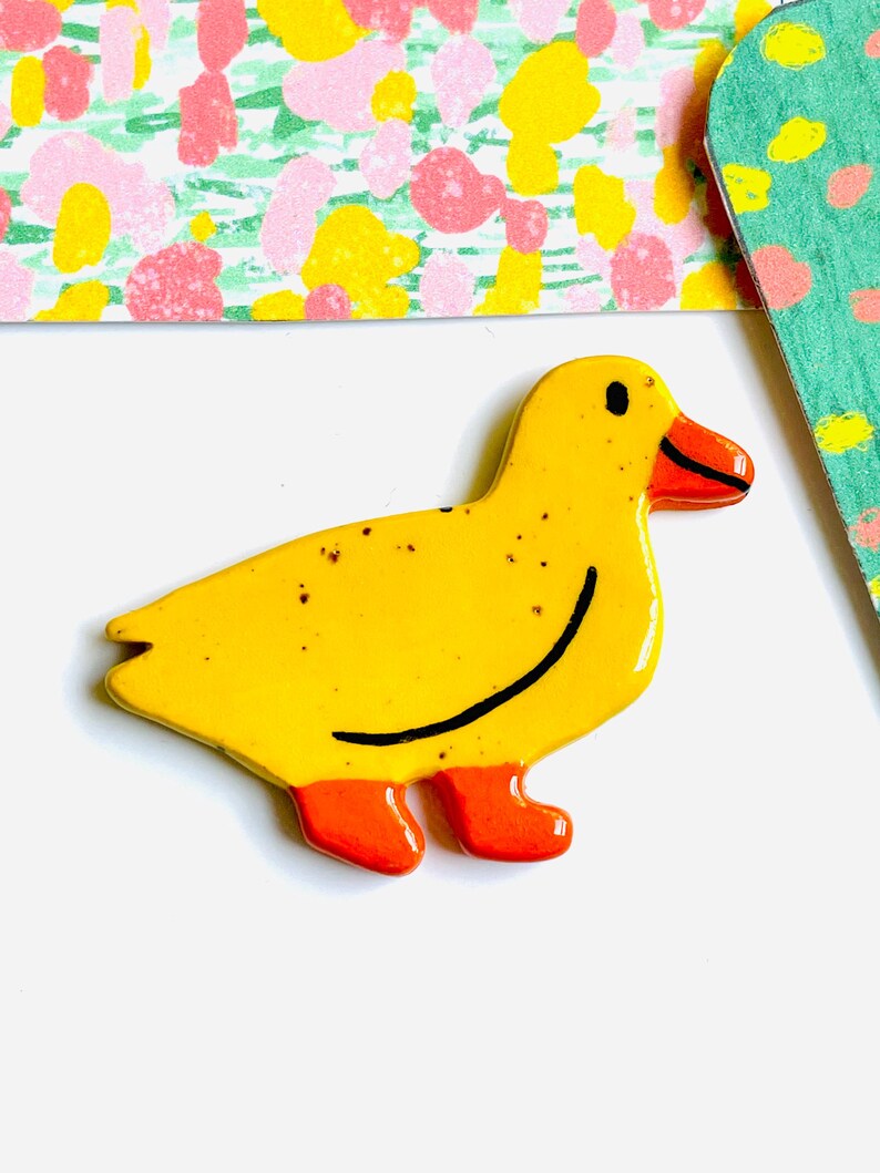 May include: A yellow ceramic duck figurine with orange feet and beak. The duck has a black smiling line and small black eyes. The background includes colorful patterned paper.
