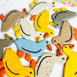 May include: A collection of ceramic duck figurines in various colors, including yellow, blue, gray, and beige. Each duck has orange feet and beaks, with black curved lines and dots for details. The ducks are arranged on a white surface.