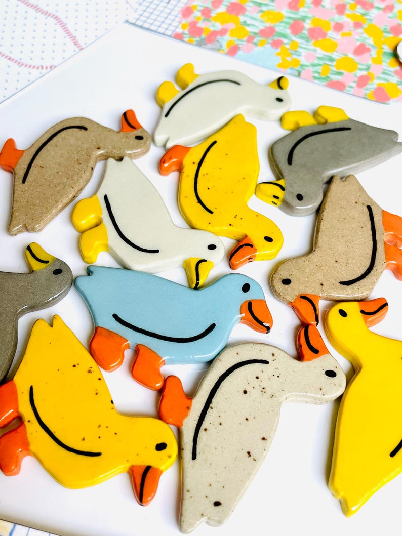 May include: A collection of ceramic duck figurines in various colors, including yellow, blue, gray, and beige. Each duck has orange feet and beaks, with black curved lines and dots for details. The ducks are arranged on a white surface.