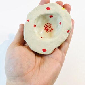 May include: A small, white ceramic dish with a red and yellow glaze. The dish has a round, indented center and is decorated with red dots. The dish is stamped with the letters "RD" in red.