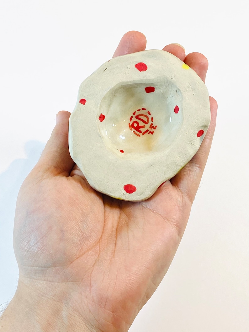 May include: A small, white ceramic dish with a red and yellow glaze. The dish has a round, indented center and is decorated with red dots. The dish is stamped with the letters "RD" in red.