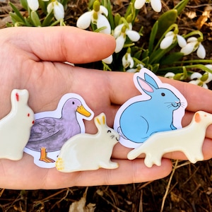 May include: A hand holding five ceramic animal figurines. The figurines are a duck, a rabbit, a rabbit, a dog, and a rabbit. The duck is purple and white, the rabbits are white, and the dog is white with brown spots.
