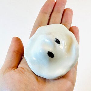 May include: White ceramic ghost figurine with black eyes and a mouth. The ghost is sitting in the palm of a hand.