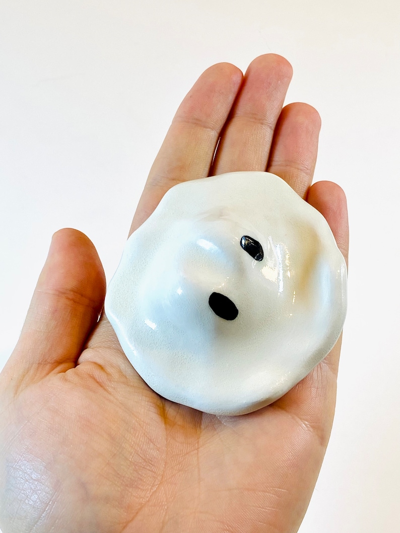 May include: White ceramic ghost figurine with black eyes and a mouth. The ghost is sitting in the palm of a hand.