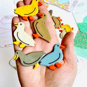 May include: A collection of colorful duck-shaped wooden pins. The pins feature various colors, including yellow, white, gray, and blue, with orange feet and beaks. The pins are held in a hand, with some cards in the background.
