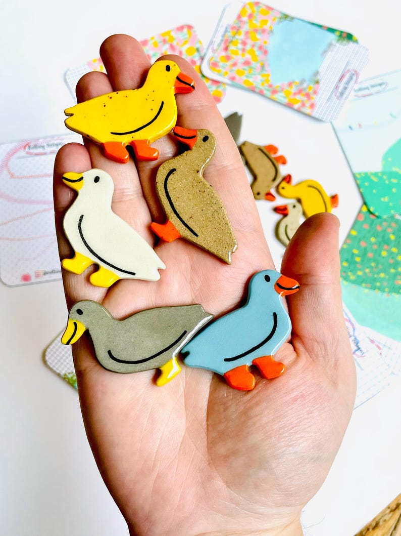 May include: A collection of colorful duck-shaped wooden pins. The pins feature various colors, including yellow, white, gray, and blue, with orange feet and beaks. The pins are held in a hand, with some cards in the background.