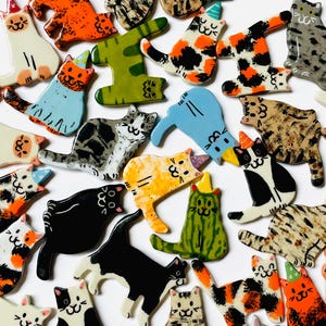 May include: A collection of flat, ceramic cat-shaped ornaments in various colors and patterns, including solid black, orange, green, and blue, as well as calico, tabby, and spotted designs. Many cats wear small, colorful party hats on their heads.