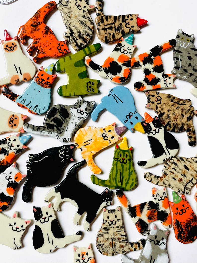 May include: A collection of flat, ceramic cat-shaped ornaments in various colors and patterns, including solid black, orange, green, and blue, as well as calico, tabby, and spotted designs. Many cats wear small, colorful party hats on their heads.