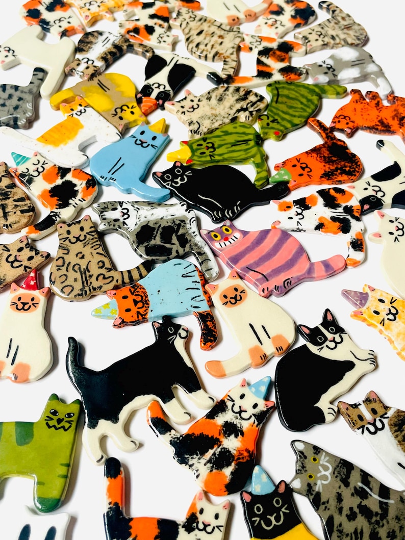 May include: A collection of numerous small, flat, ceramic cat-shaped ornaments in various colors and patterns, including calico, tabby, solid black, white, and green, with some cats wearing small, colorful party hats on their heads.