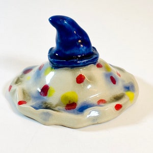 May include: A ceramic lid with a blue pointed hat on top. The lid is white with red, yellow, and blue polka dots.
