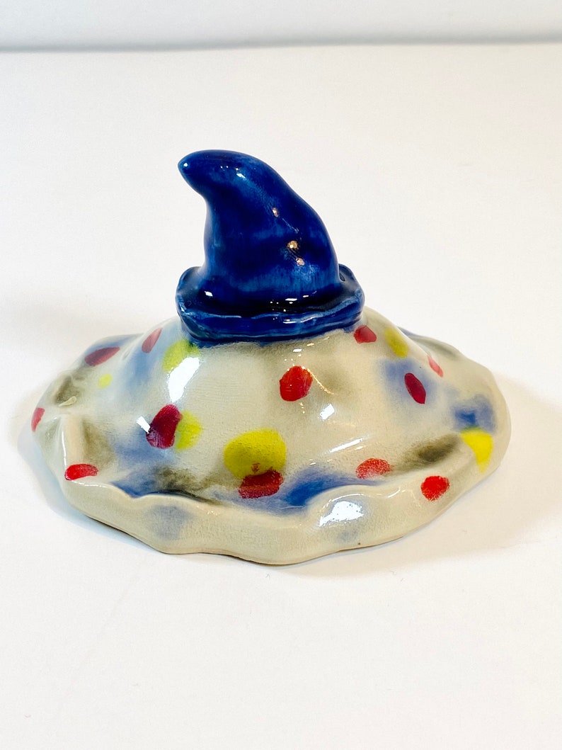 May include: A ceramic lid with a blue pointed hat on top. The lid is white with red, yellow, and blue polka dots.