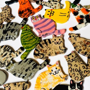 May include: A collection of flat, ceramic cat-shaped ornaments featuring various colors and patterns, including solid black, orange, yellow, green, and multi-colored tabby, calico, and spotted designs, each with hand-painted facial features.