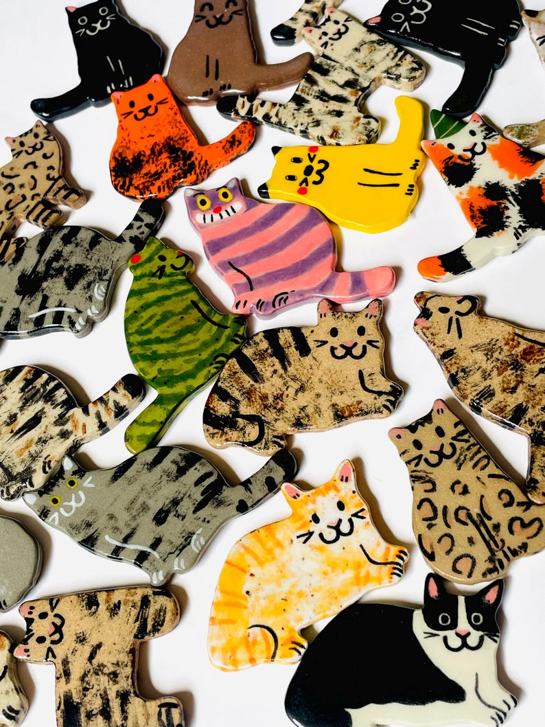 May include: A collection of flat, ceramic cat-shaped ornaments featuring various colors and patterns, including solid black, orange, yellow, green, and multi-colored tabby, calico, and spotted designs, each with hand-painted facial features.