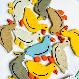 May include: A collection of ceramic duck figurines in various colors, including yellow, white, blue, and gray. Each duck has orange feet and beaks, with a black line detailing the body. The ducks are arranged on a white surface.