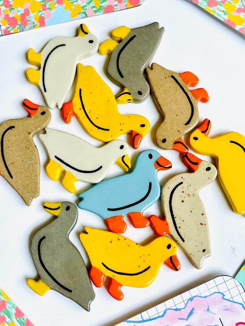 May include: A collection of ceramic duck figurines in various colors, including yellow, white, blue, and gray. Each duck has orange feet and beaks, with a black line detailing the body. The ducks are arranged on a white surface.