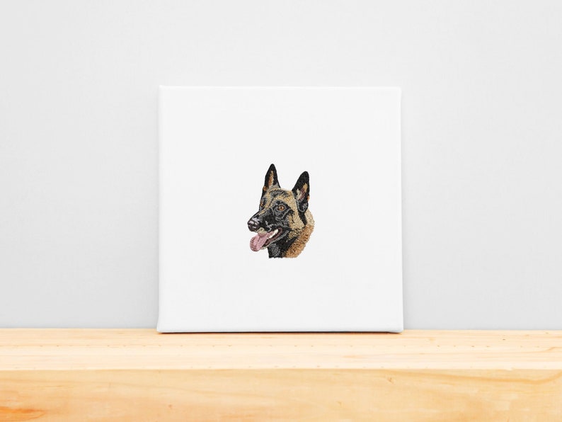 May include: A square canvas print depicting a detailed illustration of a Belgian Malinois dog's head. The dog has a black muzzle, brown and black fur, and a pink tongue. The canvas is white and rests on a wooden surface.