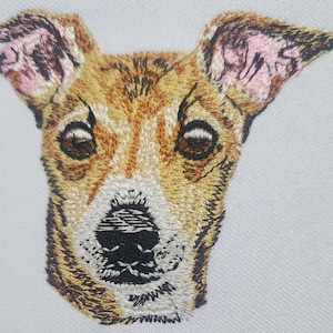 May include: Embroidered image of a dog's face. The dog has brown and white fur, with a black nose and brown eyes. The ears are pink.