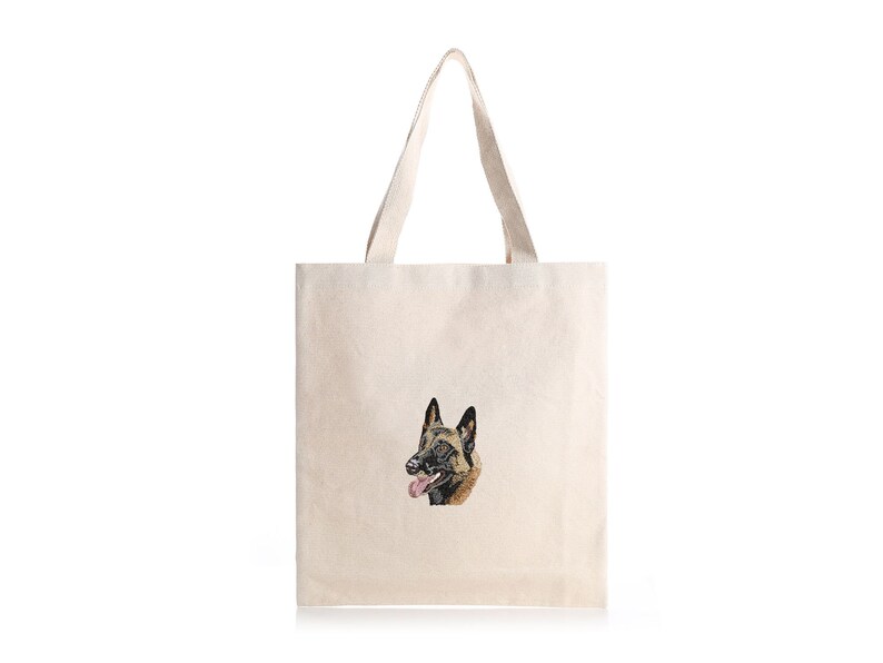 May include: A cream-coloured tote bag with an embroidered image of a Belgian Malinois dog's head. The dog has black and brown fur, a pink tongue, and is smiling. The bag has two handles and is made of a canvas-like material.