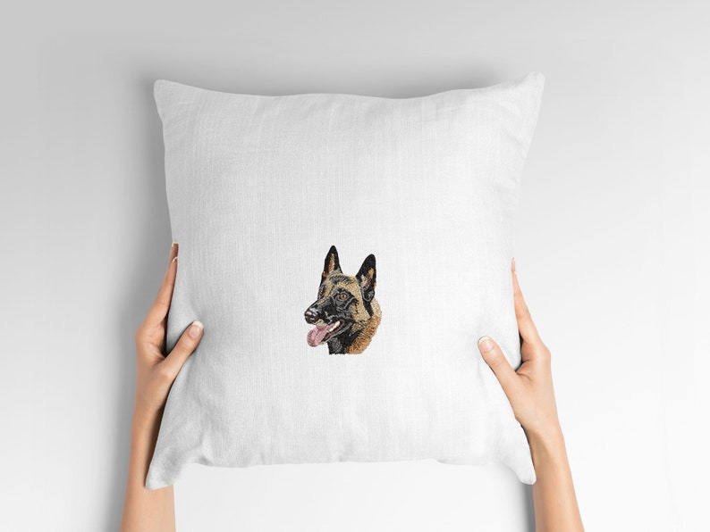 May include: A white square cushion with an embroidered image of a Belgian Malinois dog's head. The dog has brown and black fur, with its tongue visible. The cushion is held by hands against a white backdrop.