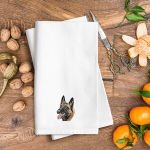 May include: White folded towel featuring an embroidered Belgian Malinois dog. The towel is placed on a wooden surface, alongside walnuts, decorative scissors, and tangerines, creating a festive atmosphere.