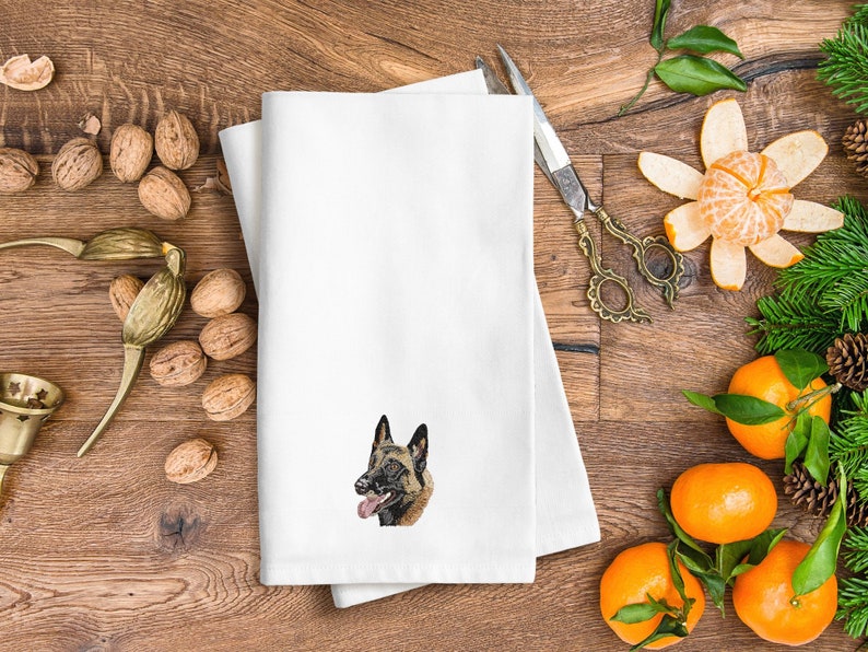 May include: White folded towel featuring an embroidered Belgian Malinois dog. The towel is placed on a wooden surface, alongside walnuts, decorative scissors, and tangerines, creating a festive atmosphere.