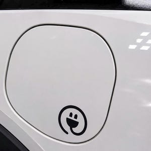 Könnte beinhalten: Eine weiße Ladeklappe für ein Fahrzeug mit einem schwarzen kreisförmigen Symbol, das einen stilisierten elektrischen Stecker zeigt. Die Ladeabdeckung ist in die glatte, glänzend weiße Karosserie des Autos integriert und wirkt sauber und modern.