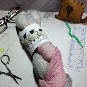 May include: A skein of yarn in shades of gray and pink, with a label that reads "Shirley String Weaving" and features three sheep.