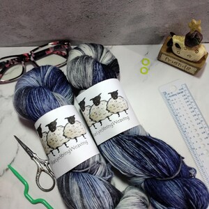 May include: Two skeins of yarn in shades of blue and grey with white flecks. The yarn is wrapped around cardboard tubes with labels that say "Marylyn String Weaving" and feature a cartoon sheep.