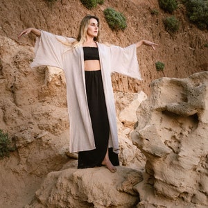 Pode incluir: Uma mulher vestindo um quimono longo bege claro com um top preto e uma saia preta está em pé em um penhasco rochoso. A mulher está olhando para a direita do quadro e tem os braços estendidos.