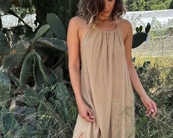 May include: A woman wearing a light brown sleeveless maxi dress with a side slit. The dress has a gathered neckline and a relaxed fit. She is standing in a field of tall grass.