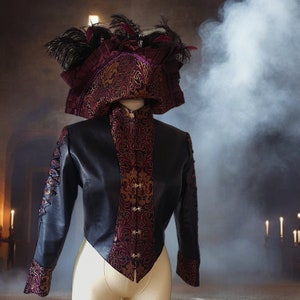 Peut inclure: Une veste noire à manches longues, dotée d'un panneau central et de poignets en damas rouge foncé et doré, agrémentée de fermoirs métalliques. La veste présente des détails à lacets sur les manches et est accompagnée d'un chapeau assorti.