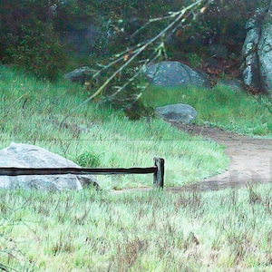 May include: A wooden fence with two posts and a horizontal rail, set in a grassy field with large rocks in the background.