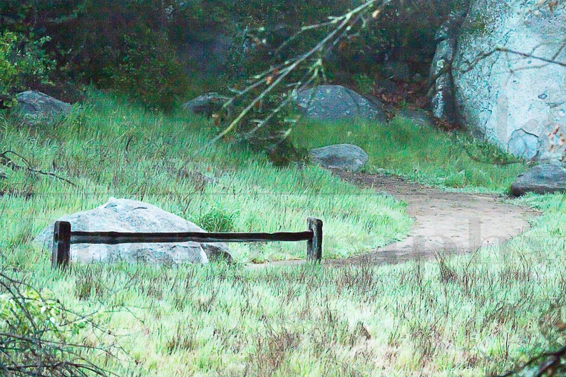 May include: A wooden fence with two posts and a horizontal rail, set in a grassy field with large rocks in the background.