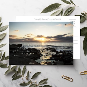 May include: A postcard with a photo of a sunset over the ocean. The sky is a mix of orange, pink, and purple. The water is a deep blue, and there are rocks in the foreground. The text on the postcard reads "La Jolla Sunset" - La Jolla, CA.