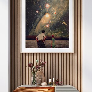 May include: A framed print of a couple standing in a field, looking up at a starry night sky with a galaxy and planets. The print is hanging on a wall with wood paneling.