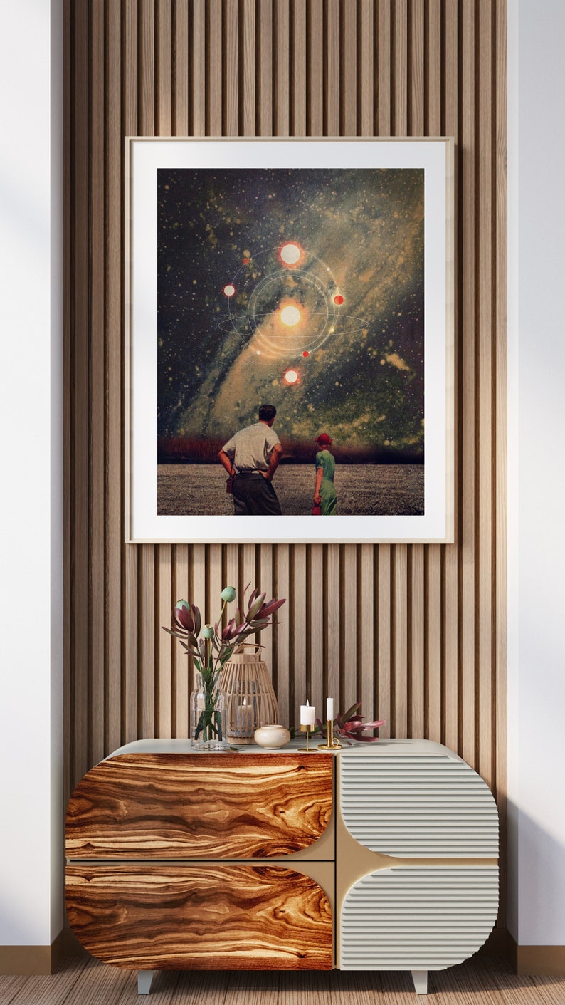 May include: A framed print of a couple standing in a field, looking up at a starry night sky with a galaxy and planets. The print is hanging on a wall with wood paneling.