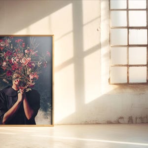 以下が含まれることがあります： 顔にピンクと赤の花束をつけた人の写真が額縁に入っています。その人は白い襟の黒いドレスを着ています。背景は濃い青です。