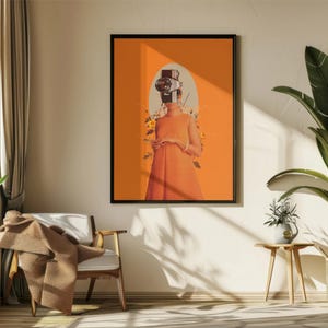 May include: A framed art print featuring a figure with a camera for a head, wearing an orange dress, set against an orange background. The artwork is displayed on a wall in a room with a window, chair, and plants.