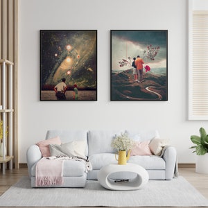 May include: Two framed art prints, one depicting a couple walking on a winding path in a landscape with a red flower, and the other depicting a starry sky with a galaxy and planets. The prints are hanging on a white wall above a light gray sectional sofa with a pink throw blanket.