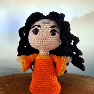 May include: A handmade crochet doll with black curly hair and large, expressive eyes. The doll wears an orange dress with yellow trim and a small blue marking on its forehead. The doll is approximately 15 cm tall.